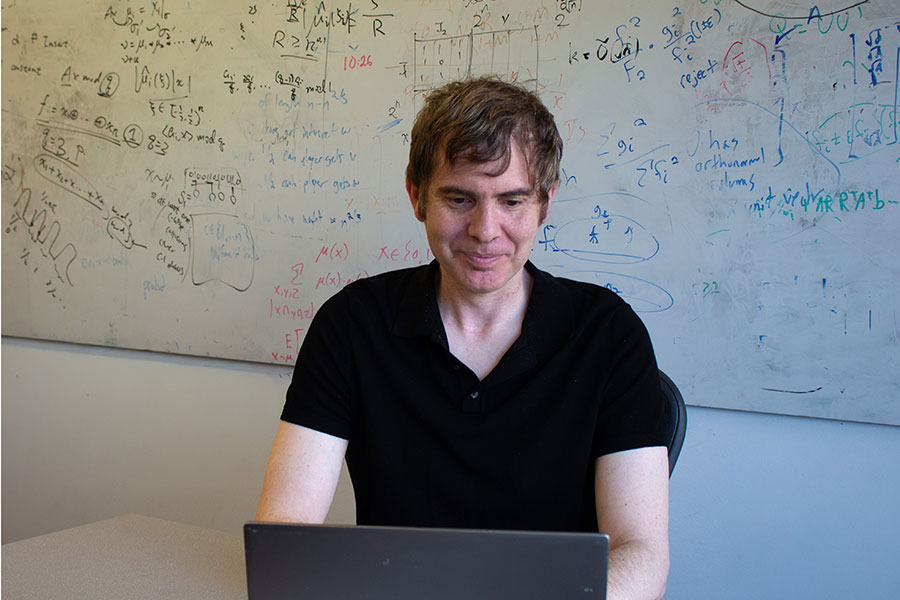 A man sits at a laptop, in front of a dry erase board covered in complicated equations.