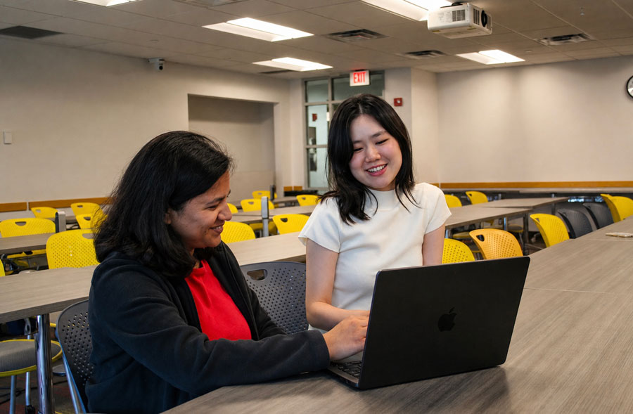 Two students sit in an empty classroom, smiling as they work on a laptop.