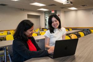 Two students sit in an empty classroom, smiling as they work on a laptop.